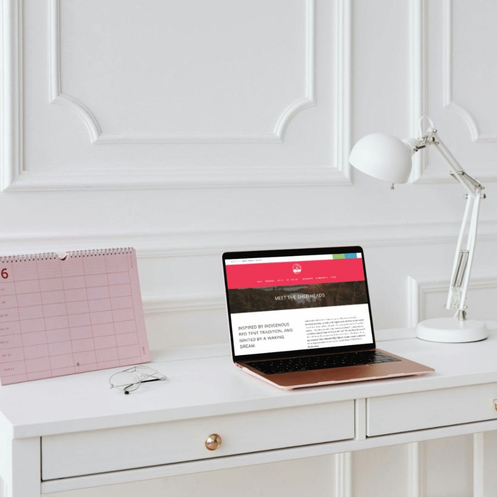 28 | Moonlit Media LLC A white desk with a pink calendar, an open laptop displaying a web designer’s site, and a white desk lamp, set against a white paneled wall.