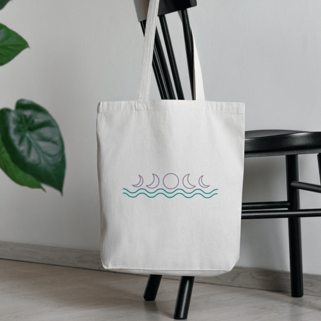 280-1 | Moonlit Media LLC A white canvas tote bag with a minimalist moon phases design by Moonlit Media hangs on the back of a black chair. A green plant is partially visible in the background, reflecting sleek style perfect for creative logo design services.