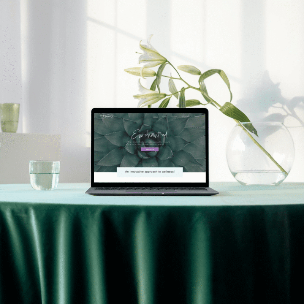 281-1 | Moonlit Media LLC A laptop on a green table displays a custom website design for a wellness site. Next to it are a glass of water, an empty round vase, and white lilies in the background, all lit by natural sunlight.