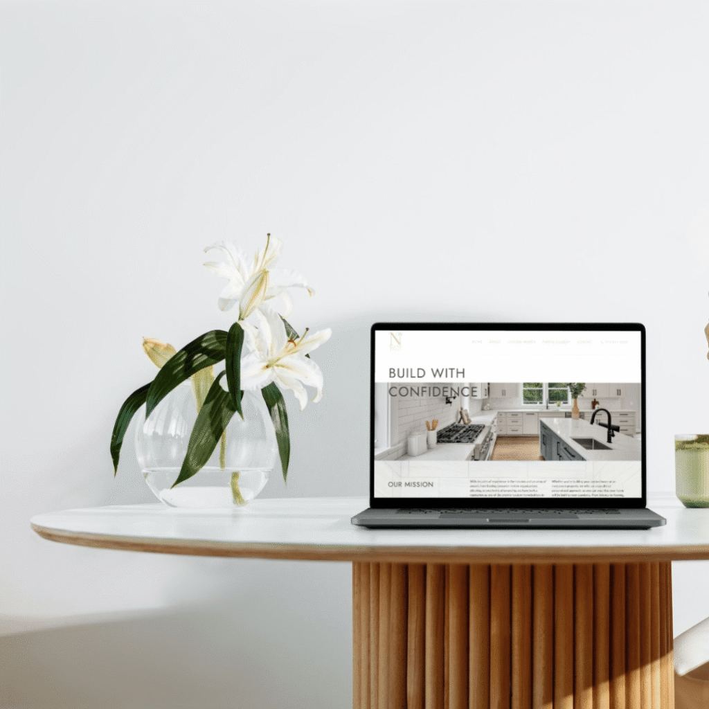 A laptop displaying a website sits on a round white table with a wooden base. Next to it is a glass vase with white lilies and green leaves—an inviting space perfect for exploring web design services. The background is a plain white wall.