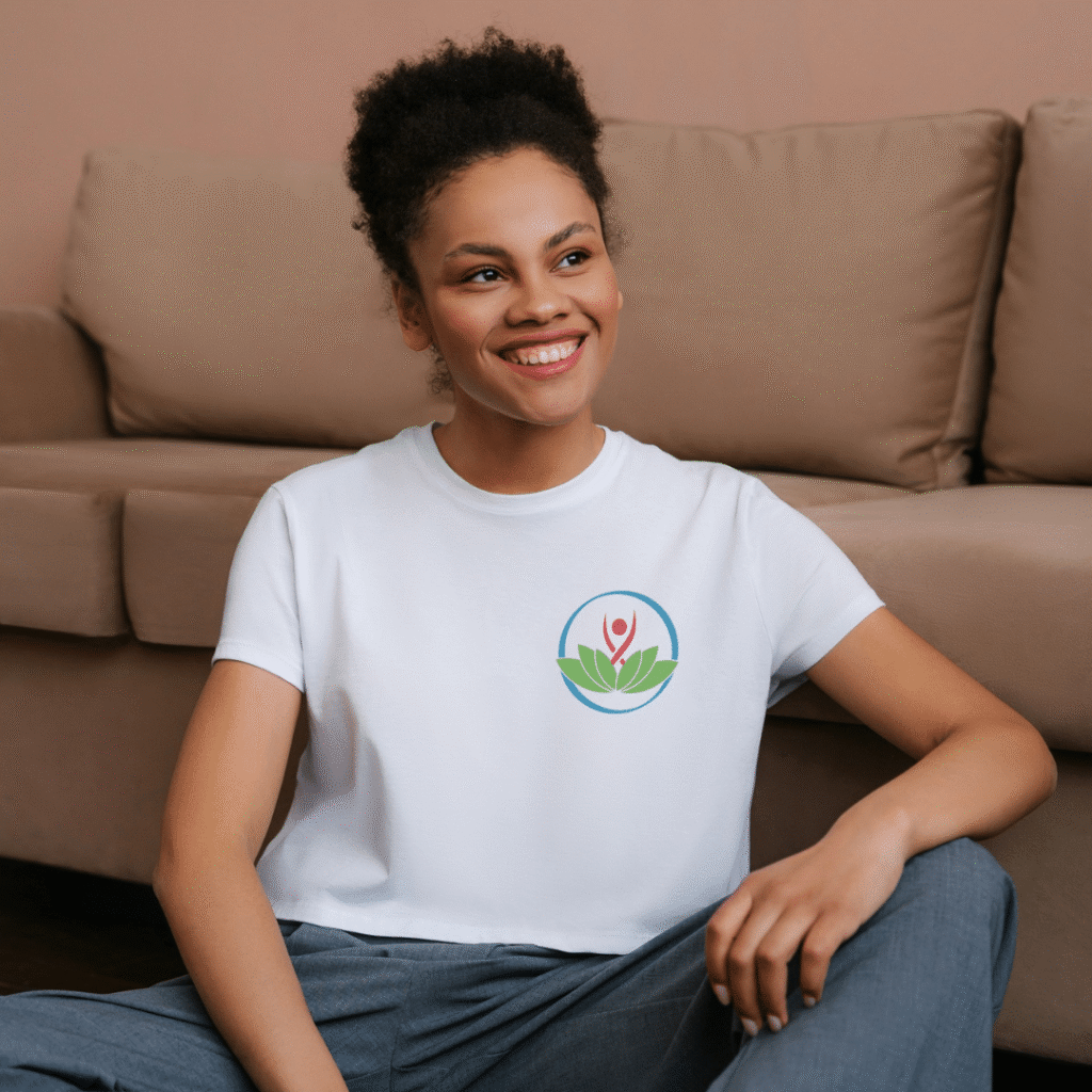 29 | Moonlit Media LLC Smiling woman with short curly hair sits on the floor in front of a beige couch, wearing a white t-shirt featuring a colorful custom logo design of a person and leaves.