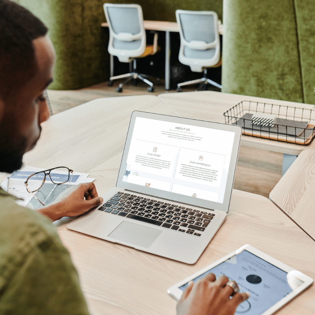 A person sits at a desk in a modern office, working on a laptop displaying an About Us webpage for Moonlit Media, known for custom logo design. A tablet and glasses rest on the desk, with empty chairs and green dividers in the background.