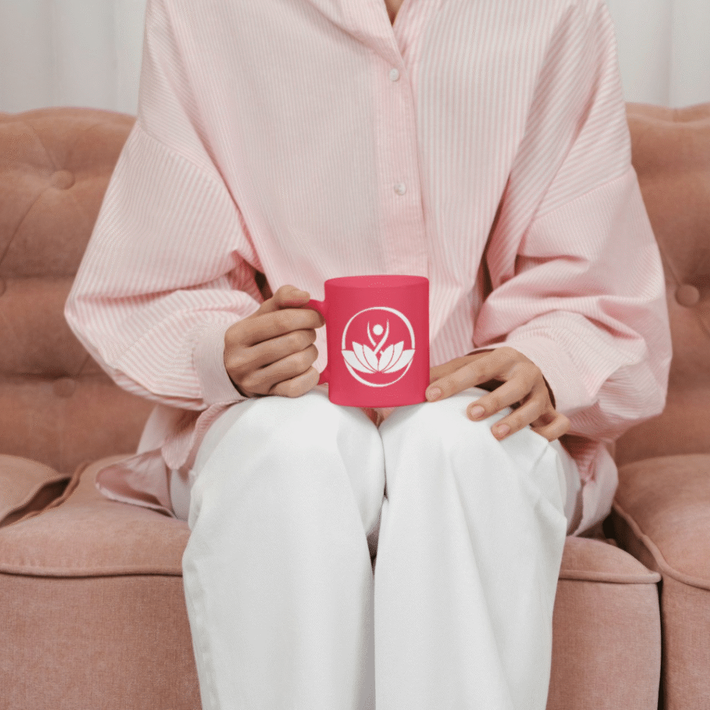 30 | Moonlit Media LLC A person in a light pink shirt and white pants sits on a pink sofa, holding a bright pink mug featuring a custom logo design with a white lotus flower and human figure. The person’s face is not visible.