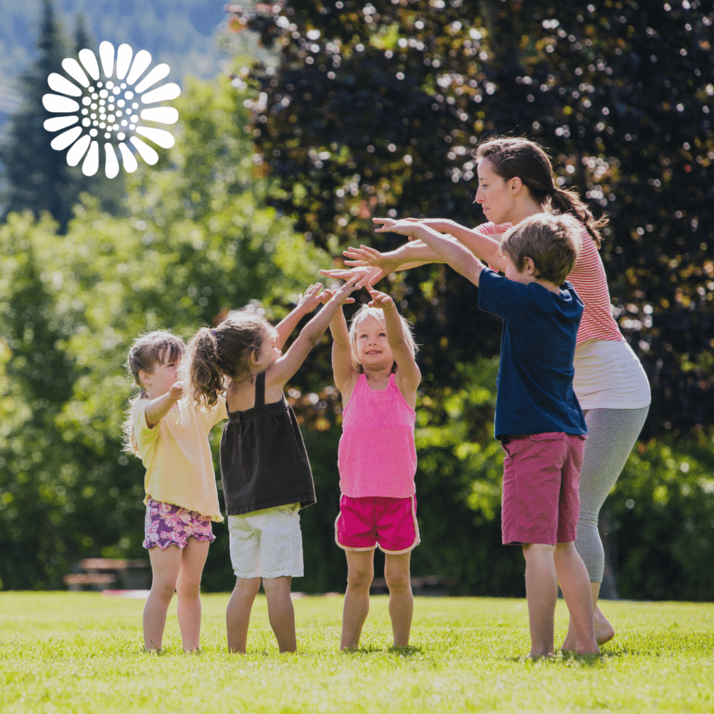 A woman and four young children stand in a grassy field, holding hands in a circle and raising their arms together. The background shows trees and sunlight, with a custom logo design by Moonlit Media in the top left corner.