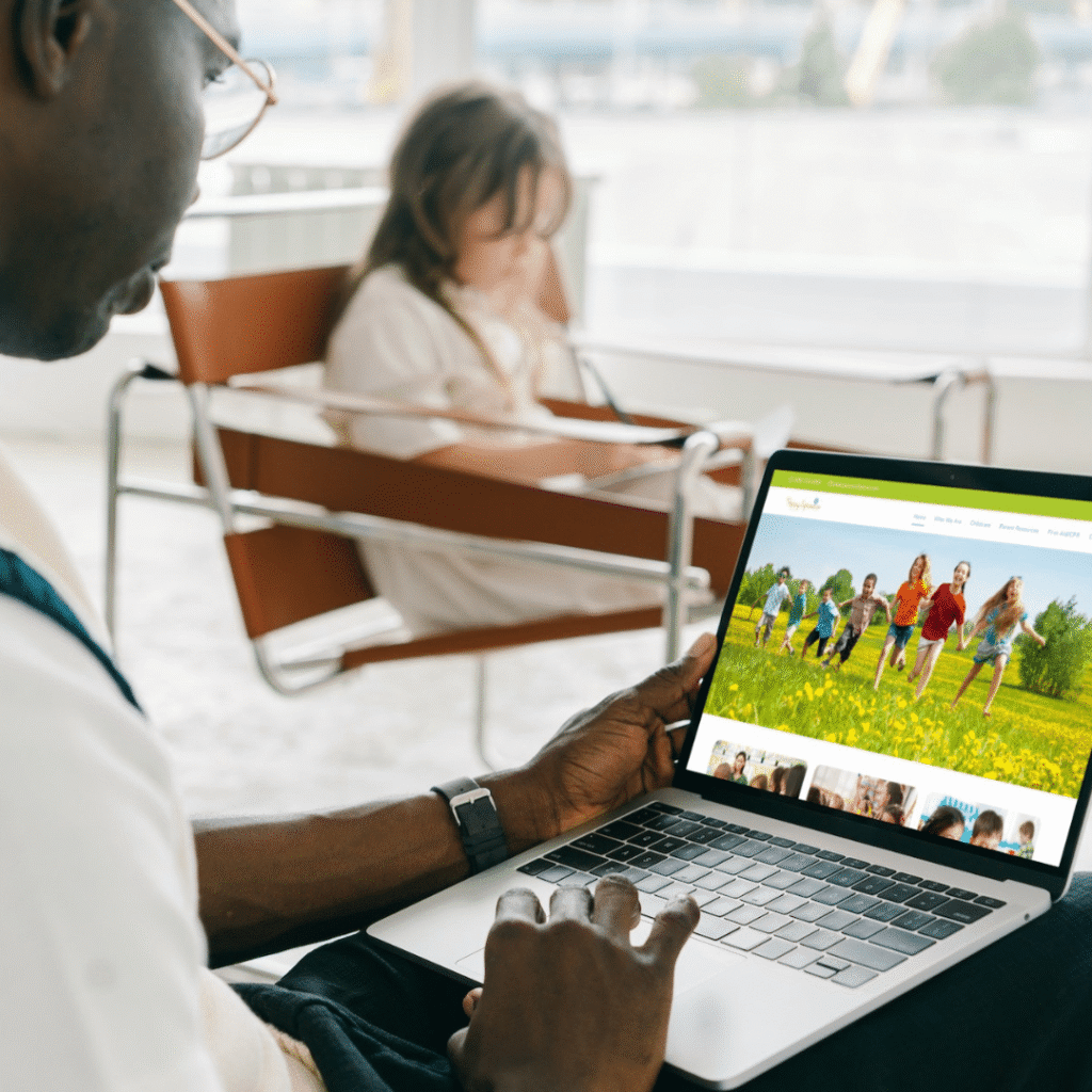 A person using a laptop with a custom website design showing children running in a field, while a child sits reading in the background on a modern chair in a bright room.