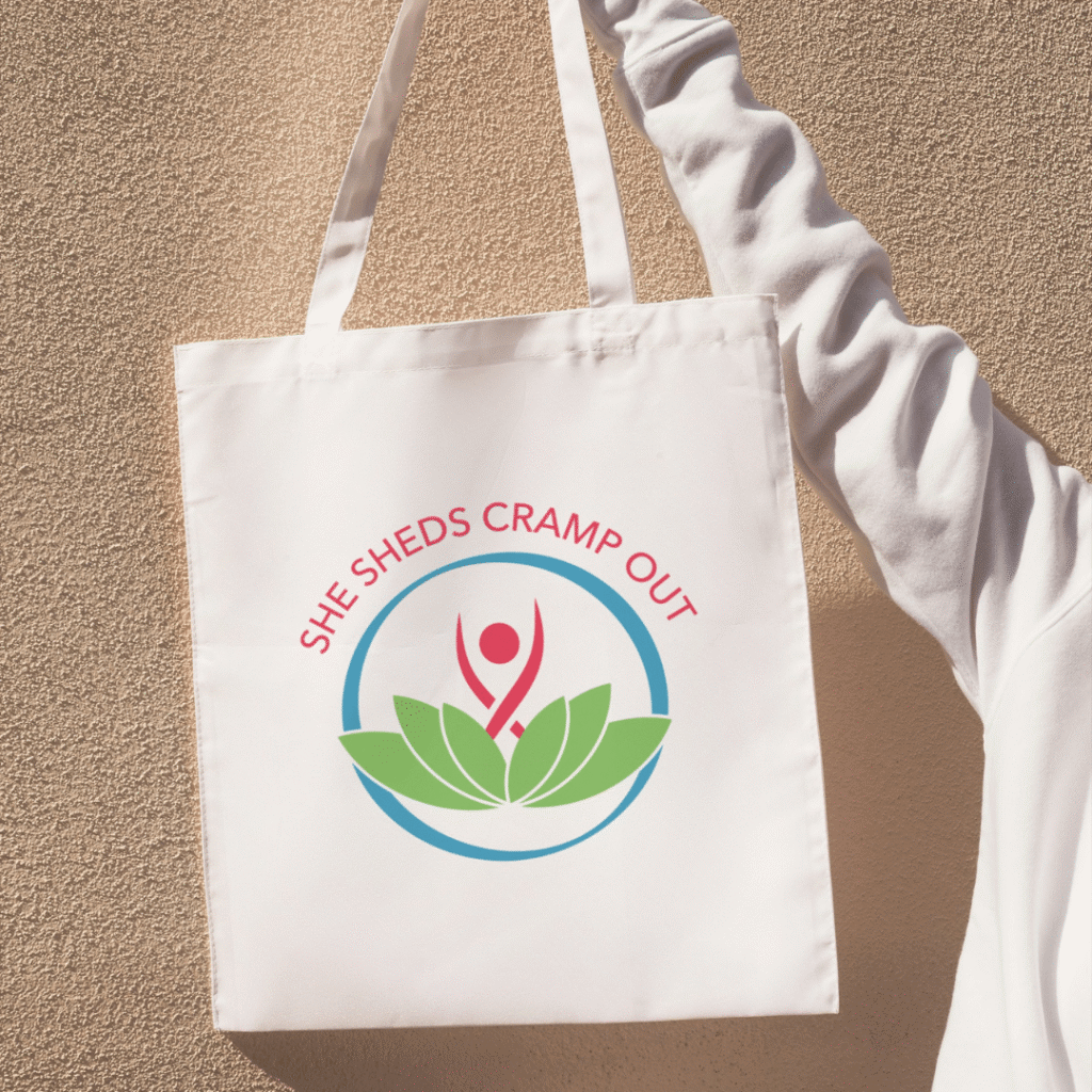 31 | Moonlit Media LLC A person in a white sleeve holds a white tote bag with a custom logo design—a stylized figure, green leaves, and SHE SHEDS CRAMP OUT in red—against a textured wall. Perfect for web designers seeking creative inspiration.