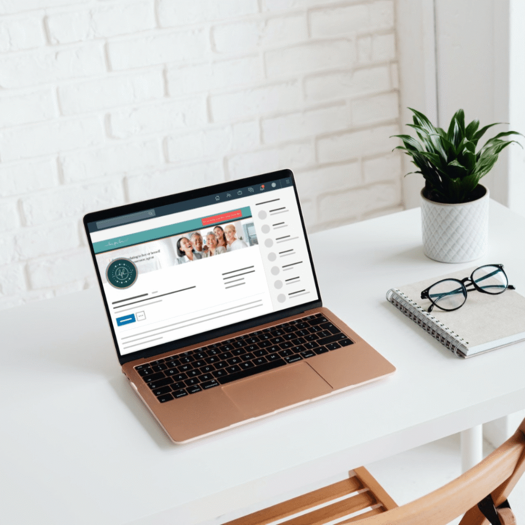 A rose gold laptop displaying a custom website design sits on a white desk beside a potted plant, eyeglasses, and a spiral notebook, with a white brick wall in the background.