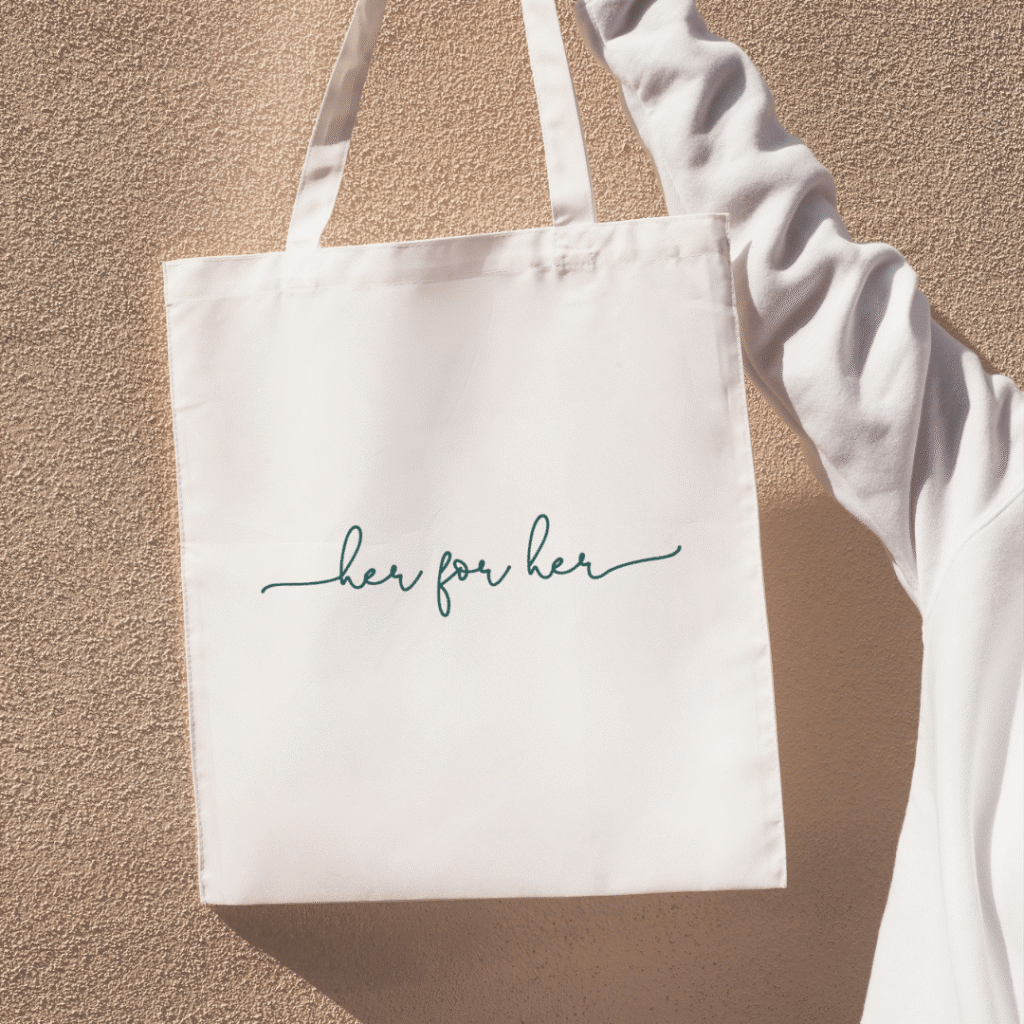 A person wearing a white long-sleeve shirt holds a white tote bag with handwritten-style green text that reads her for her, against a textured beige wall—perfect inspiration for custom logo design or creative branding ideas.