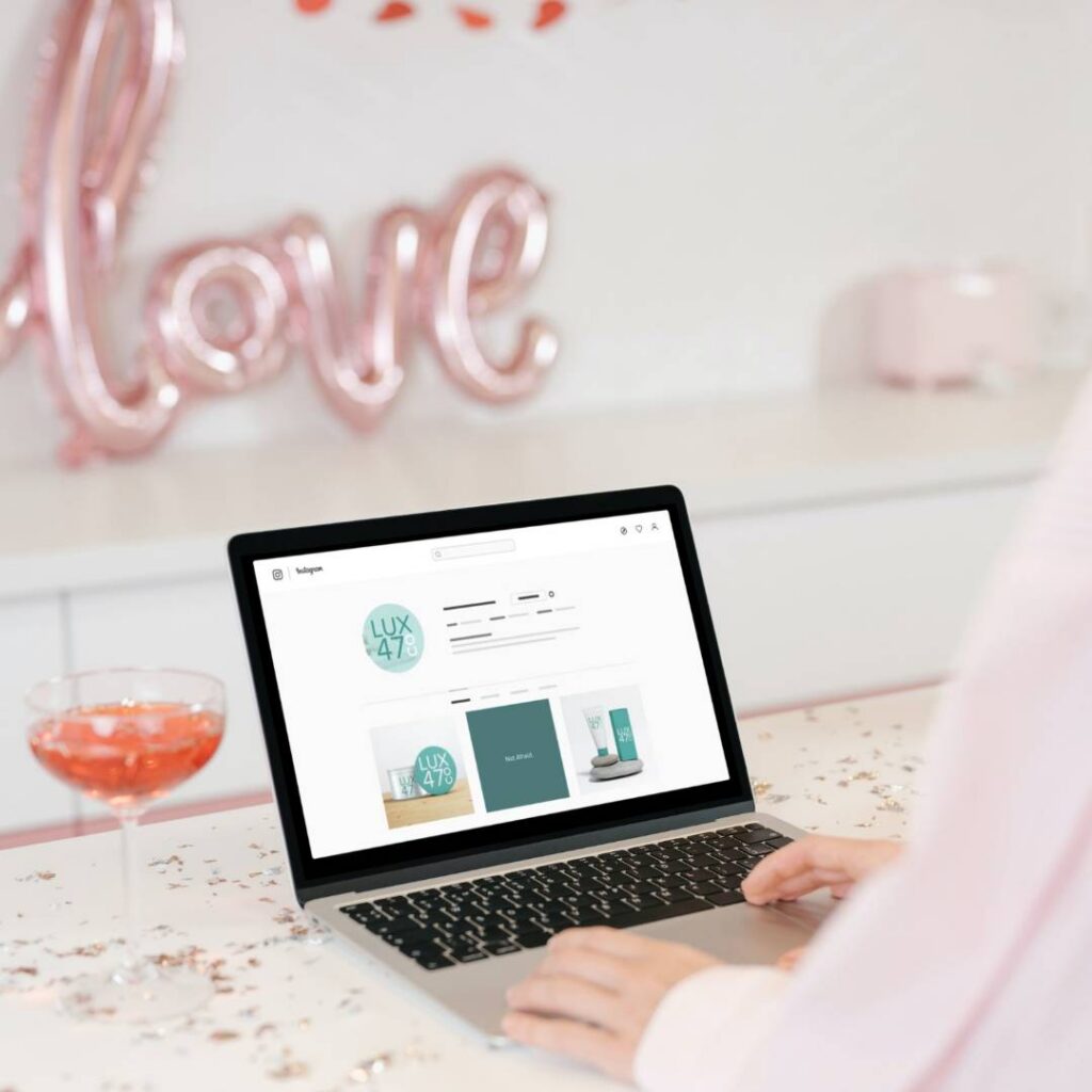 | Moonlit Media LLC A person uses a laptop on a white table decorated with confetti, a pink drink, and a love balloon in the background. The laptop screen displays a custom website design with various product images.
