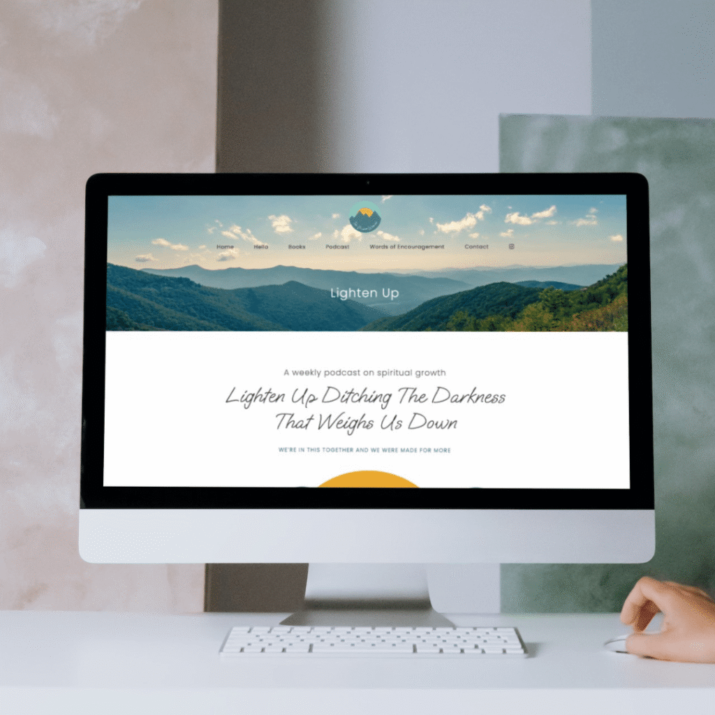 A hand rests on a desk next to an iMac displaying a website for the Lighten Up podcast, featuring moonlit media-inspired web design services and a mountain landscape banner with the tagline Ditching The Darkness That Weighs Us Down.