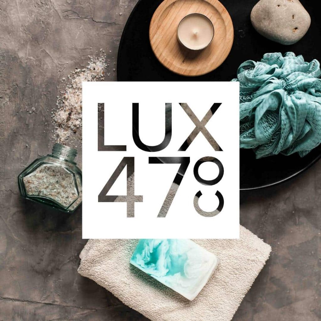 Flat lay of spa items on a gray surface, including a candle, bath salts, pumice stone, blue bath pouf, towel, and marbled soap. Perfect inspiration for web design services. The text LUX 478 is overlaid in bold letters.