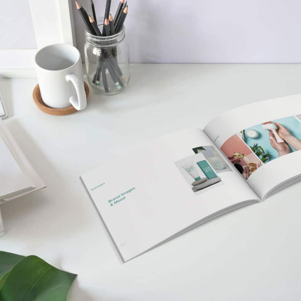 | Moonlit Media LLC An open magazine showcasing brand images lies on a white desk beside a cup, pencils, a notebook, and a green leaf—reflecting the clean, minimalistic aesthetic that Moonlit Media brings to custom website design and logo design services.