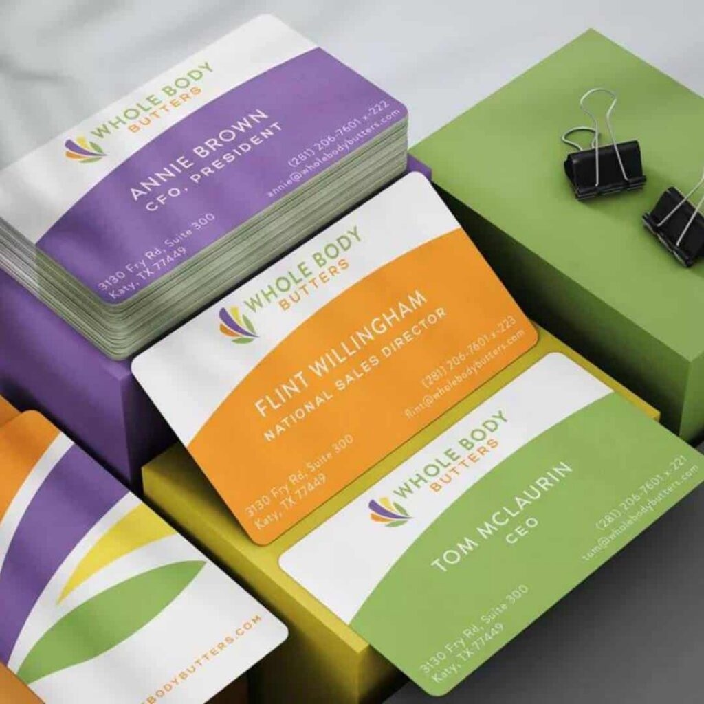 A stack of colorful business cards for Whole Body Butters with names, titles, and contact info—featuring purple, orange, and green designs—showcases moonlit media’s custom website design. Two binder clips are nearby on a gray surface.