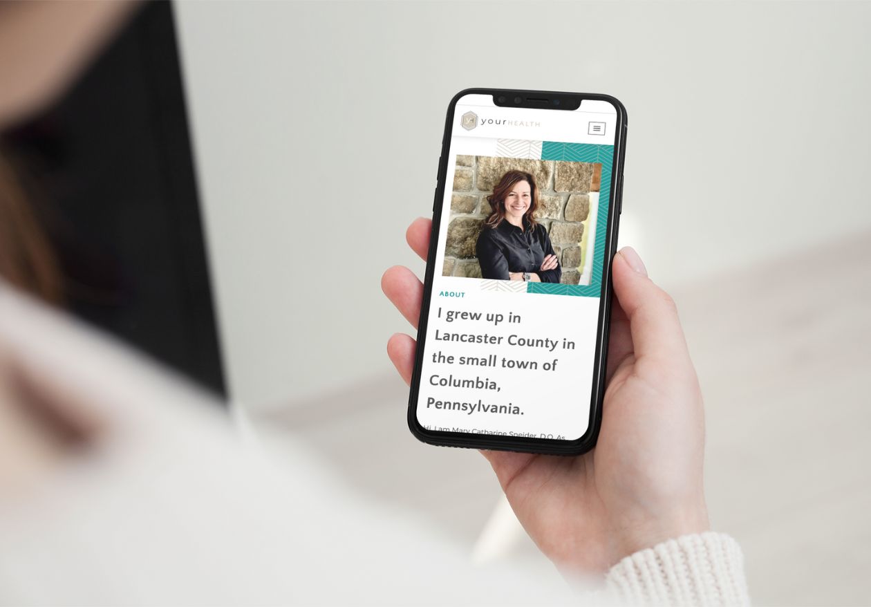 A person holds a smartphone displaying a web designer’s profile from Moonlit Media—a smiling woman with curly hair stands before a stone wall. The text highlights her roots in Lancaster County, Columbia, Pennsylvania.