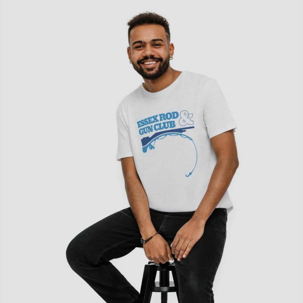 A smiling man with a beard sits on a black stool, wearing a white t-shirt with a blue graphic that reads Asset Rod & Gun Club. He has dark hair, black jeans, and looks ready to chat about web design services or Moonlit Media.