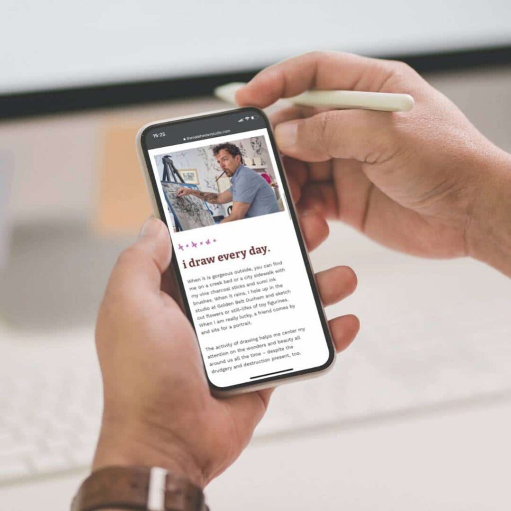 A person holds a smartphone displaying an article titled “i draw every day,” featuring a photo of a man painting on a canvas. The web designer at the desk uses a stylus, with a computer keyboard nearby.