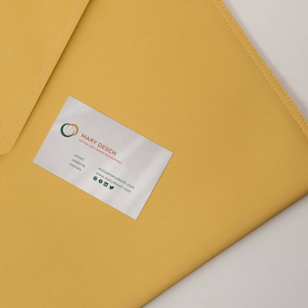 | Moonlit Media LLC A business card for Mary Desch, a web designer at Moonlit Media, rests on a mustard yellow folder. The card features contact details, a logo with intertwined circles, and social media icons on a light, neutral background.