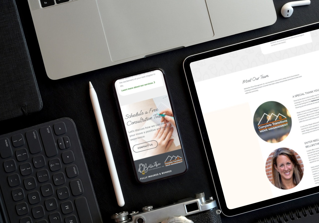 A smartphone, tablet, keyboard, and laptop are arranged on a dark desk. The phone displays a consultation offer for logo design services, while the tablet shows a “Meet Our Team” page featuring a web designer’s photo. Part of a camera is also visible.