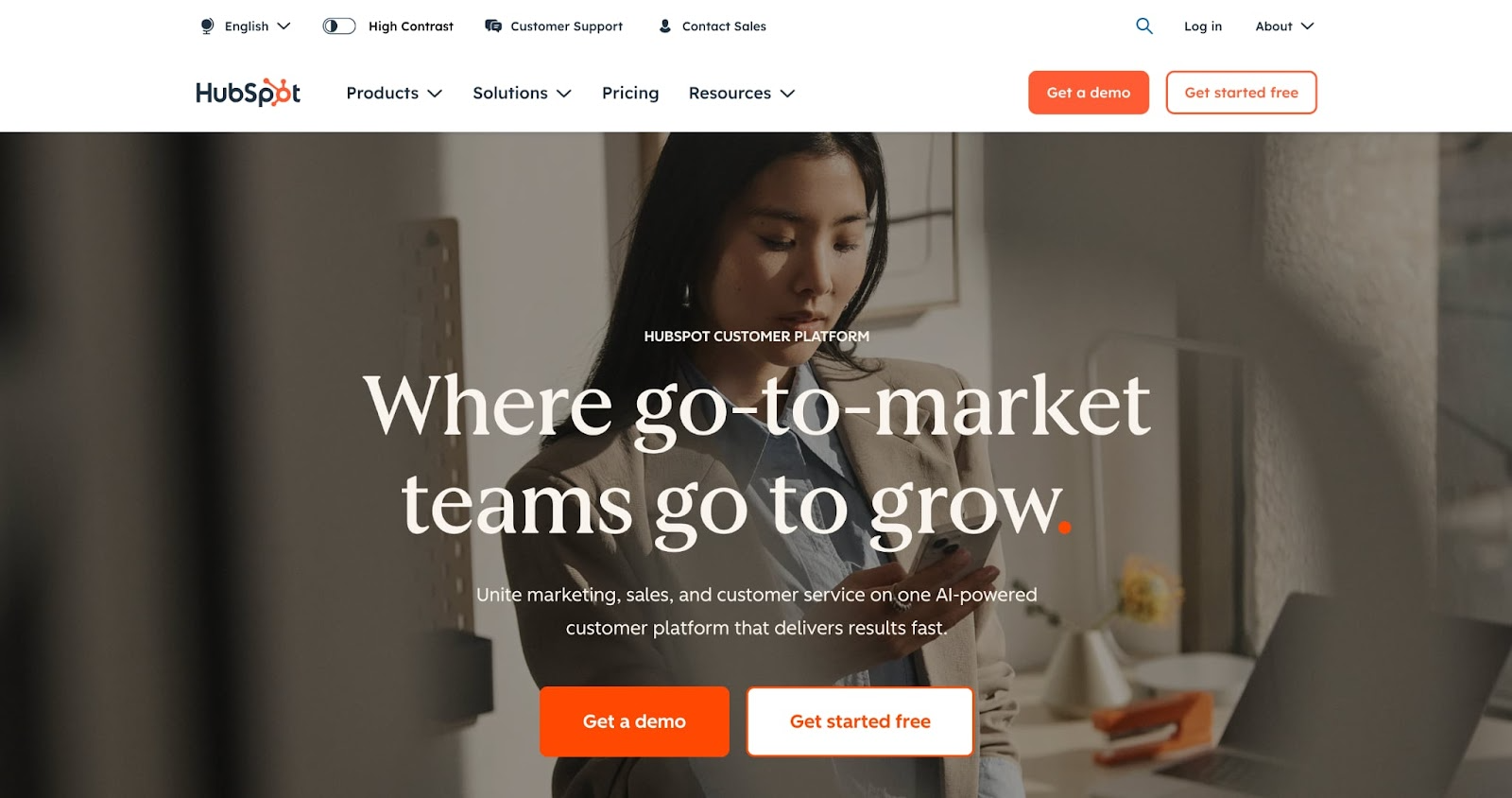 A woman stands at a desk using her phone and laptop. The HubSpot website banner displays the text, Where go-to-market teams go to grow, with navigation menus and orange buttons for demos, starting for free, and logo design services.