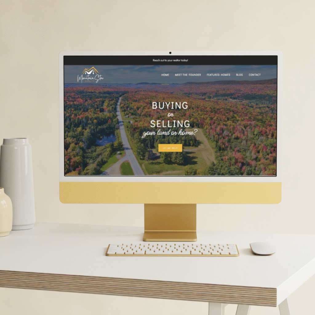 A modern computer on a beige desk displays a real estate website, crafted by a web designer, with an aerial forest view and text offering to buy or sell land or homes. A keyboard, mouse, mug, and vase complete the workspace.