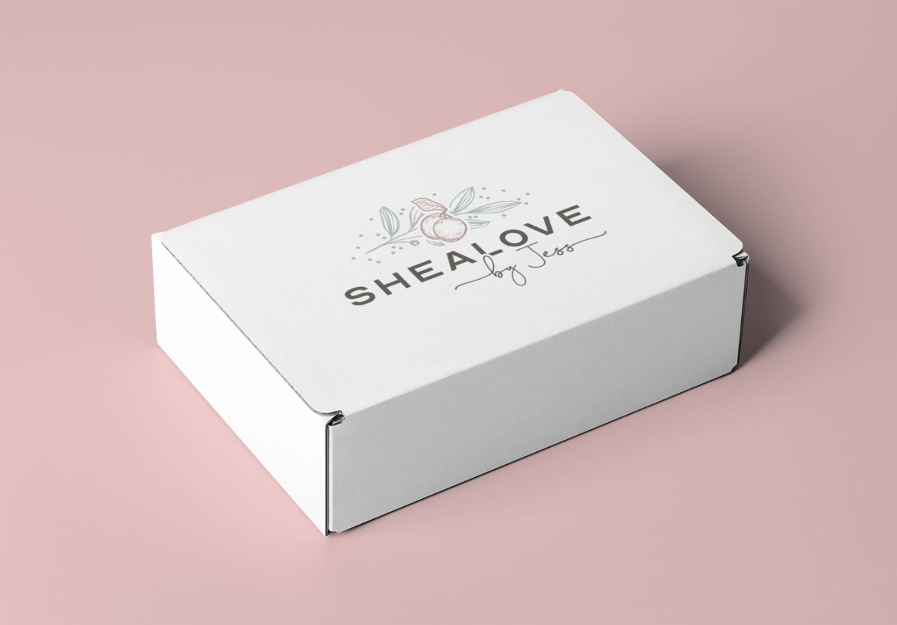 A white rectangular box on a pink surface displays a custom logo design with a stylized shea nut and leaves, and the text “SHEALOVE by Jess” on the lid, crafted by web designer Moonlit Media.