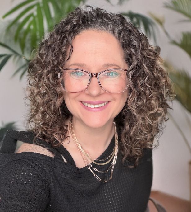 A woman with curly brown hair and glasses smiles at the camera. She is wearing a black top and layered gold necklaces—an inspiring face behind a woman-owned web design brand. Green leafy plants blur softly in the background.