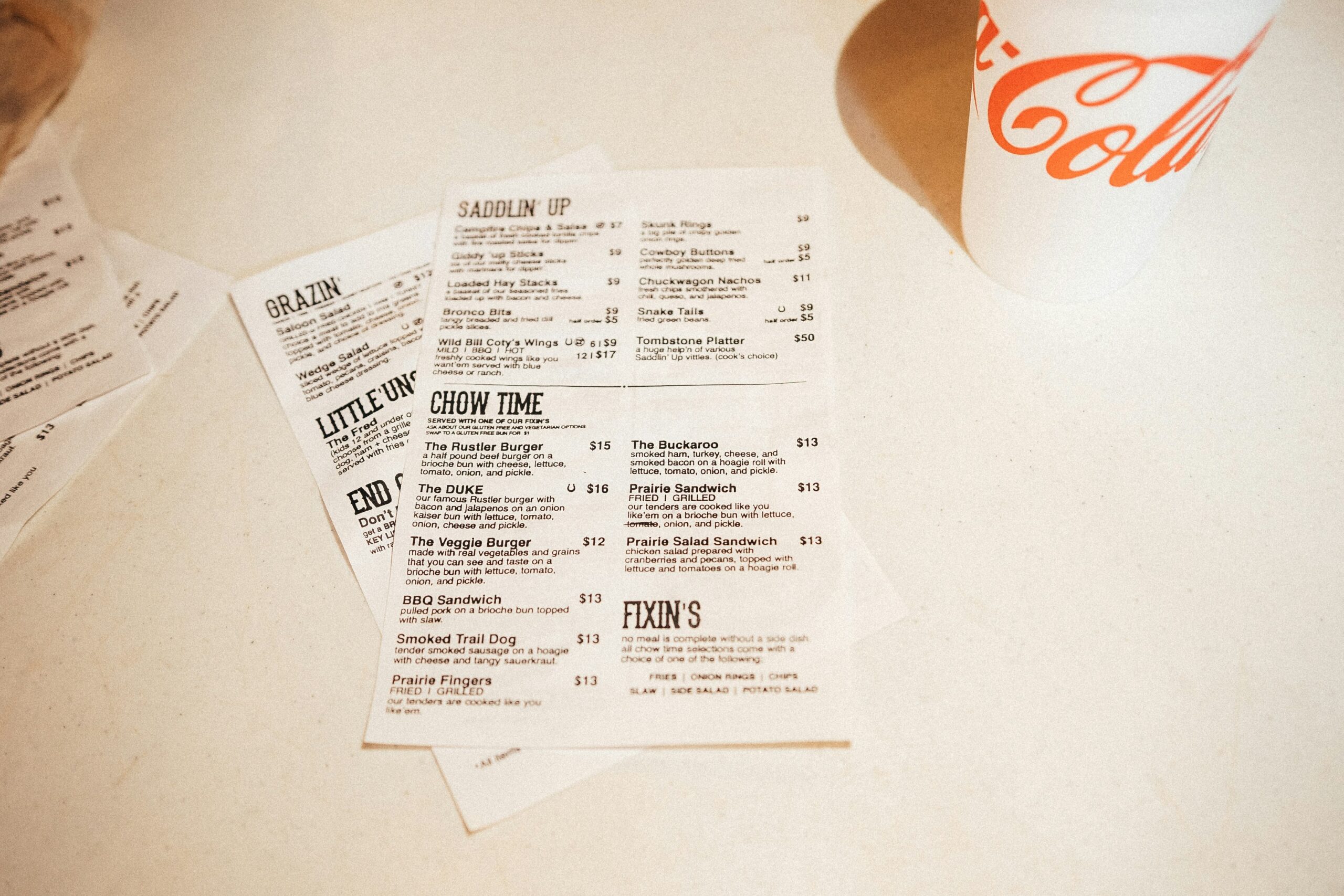 A close-up of a restaurant menu with burgers, sandwiches, wings, and sides next to a Coca-Cola cup. The menu highlights the eatery's unique branding, enhanced by professional logo design services.