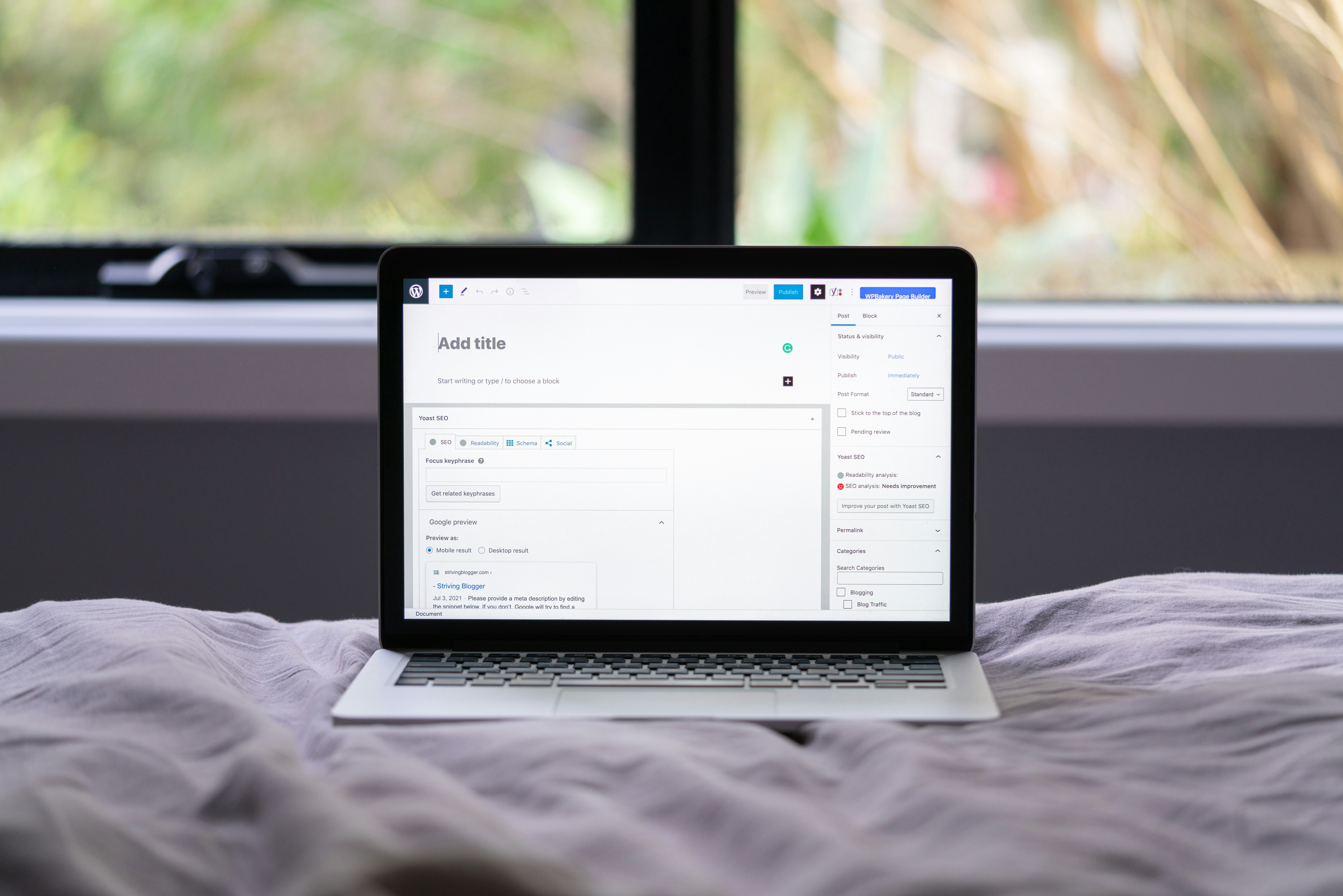 A laptop sits on a bed with a WordPress post editor open on the screen, in front of a window showing greenery—perfect for working on web design services or crafting a custom logo design.