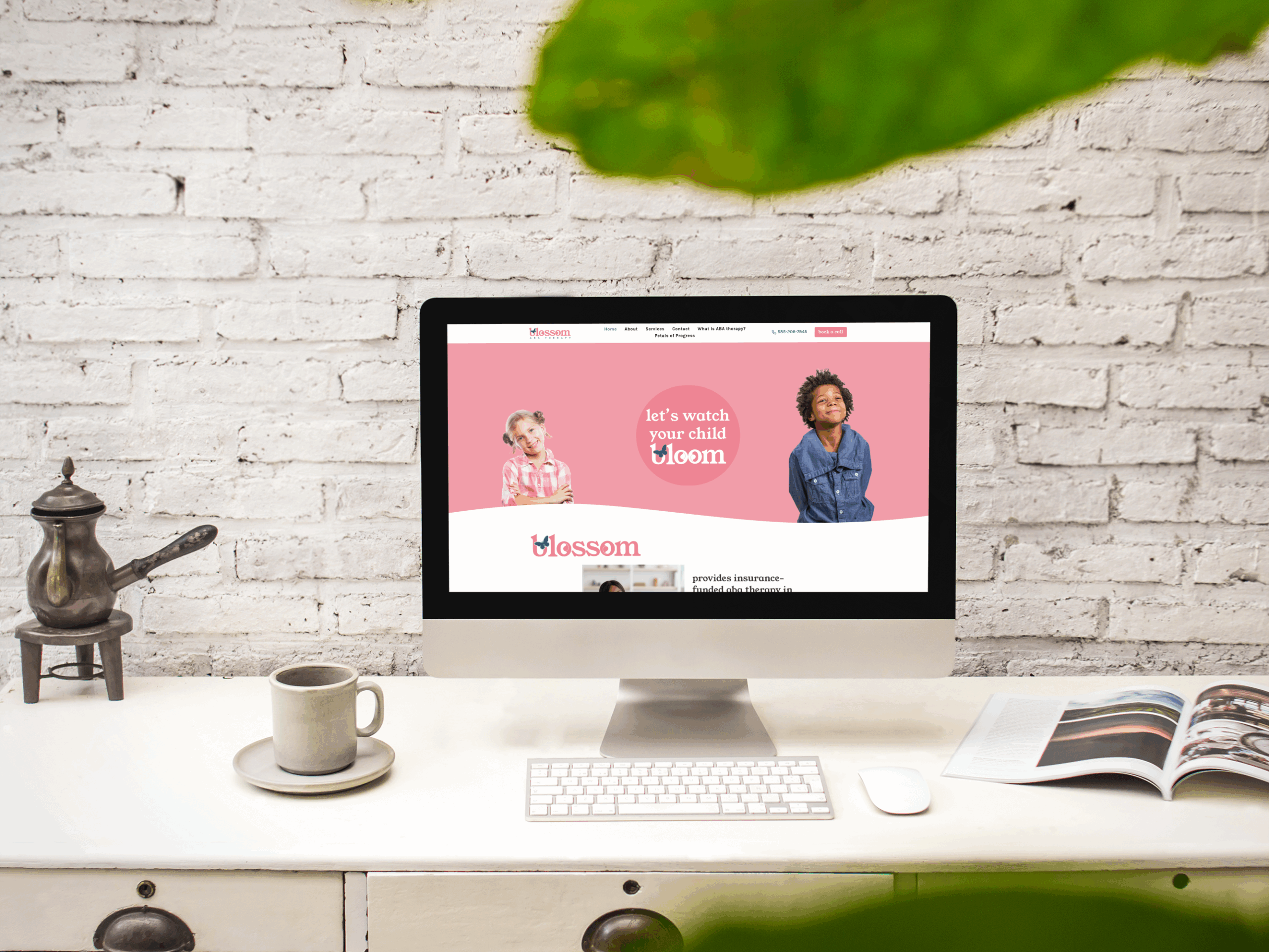 A desktop computer on a white desk displays a children's website with pink background. The scene, perfect for a web designer, includes a keyboard, mug, teapot, open magazine, and plant leaf against a white brick wall.