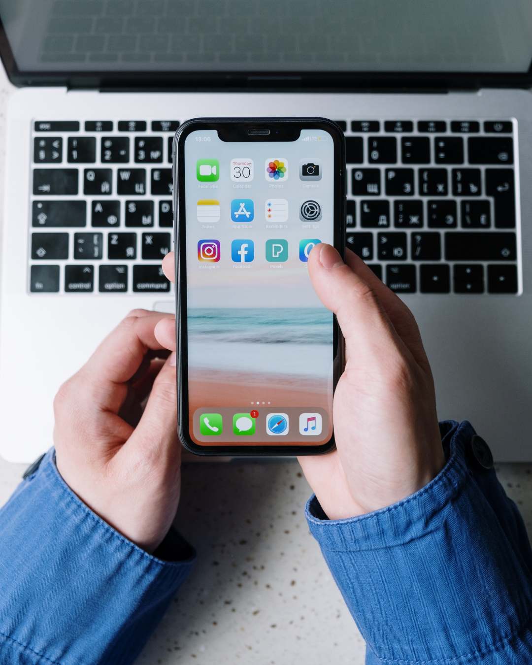 a picture of a person's hands holding a phone with the Home Screen of an iPhone with a computer in the background