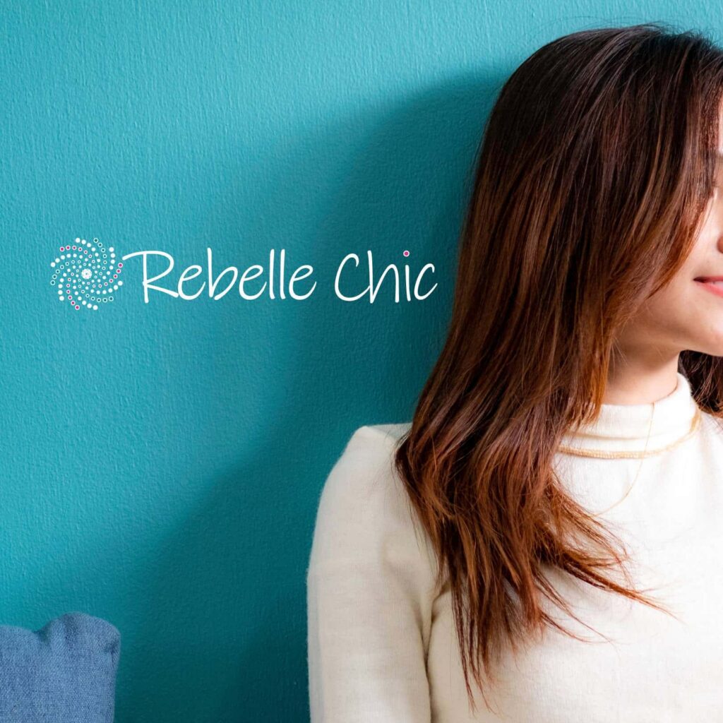A woman with long brown hair and a white sweater sits against a teal wall featuring the Rebelle Chic logo, showcasing custom website design. Her face is partially out of frame, and she is smiling slightly beside the dotted swirl design.