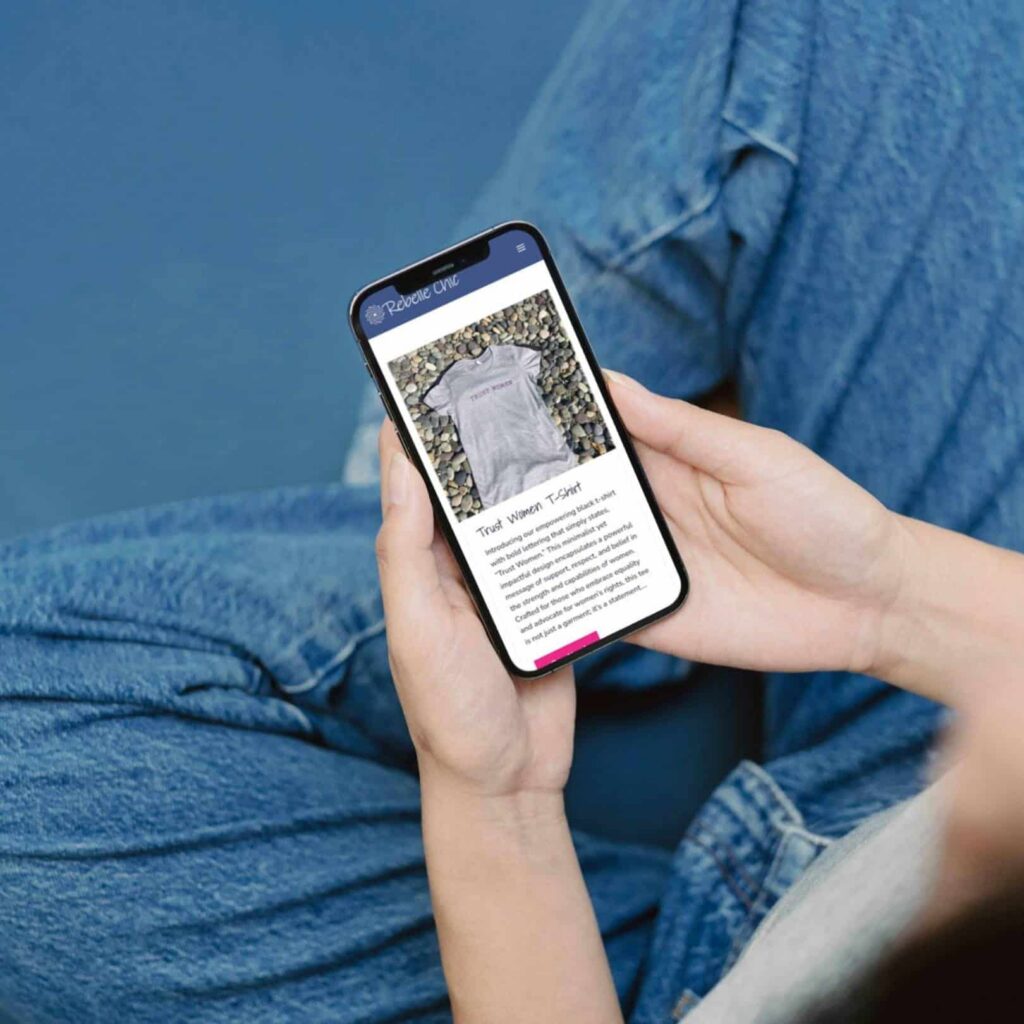 A person in blue jeans sits cross-legged, holding a smartphone displaying an image and description of a gray t-shirt on Moonlit Media’s web design services website.
