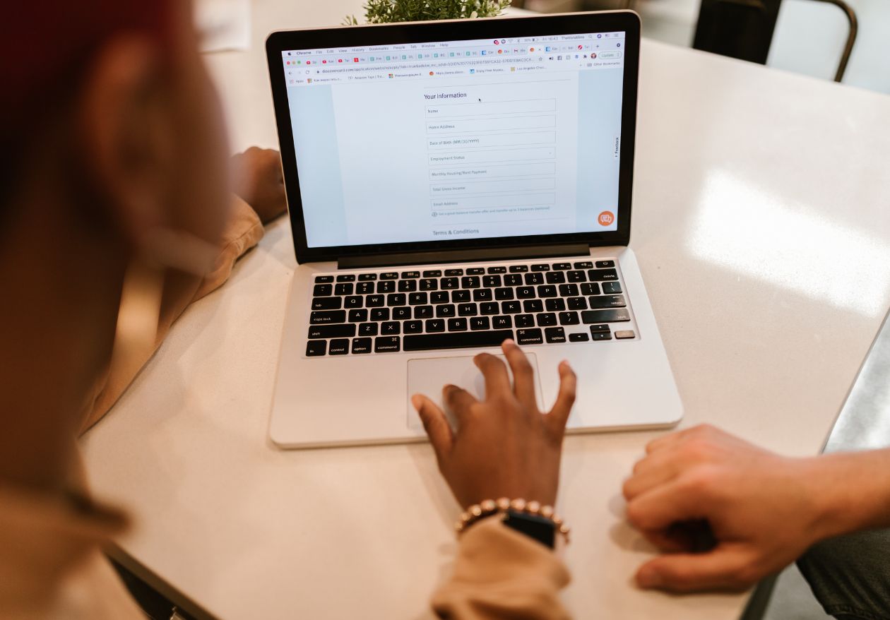 Two people sit at a table using a laptop. One person's hand is on the keyboard, and the screen displays an online form for custom website design, featuring various fields to fill out.