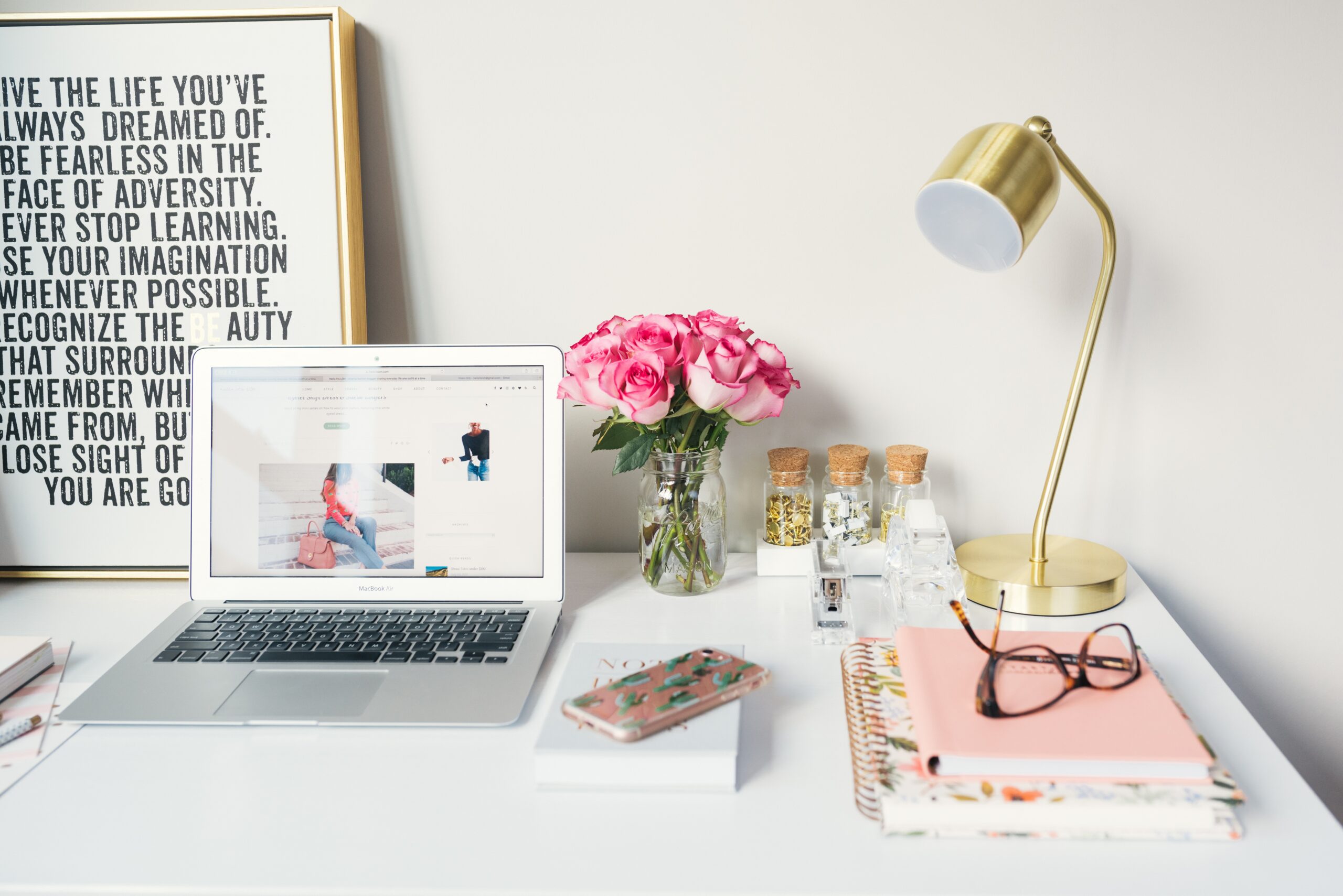 A tidy desk with a laptop, pink flowers in a vase, a smartphone, notebooks, glasses, a gold desk lamp, and a framed motivational quote—perfect inspiration for any web designer working on custom website design projects at Moonlit Media.