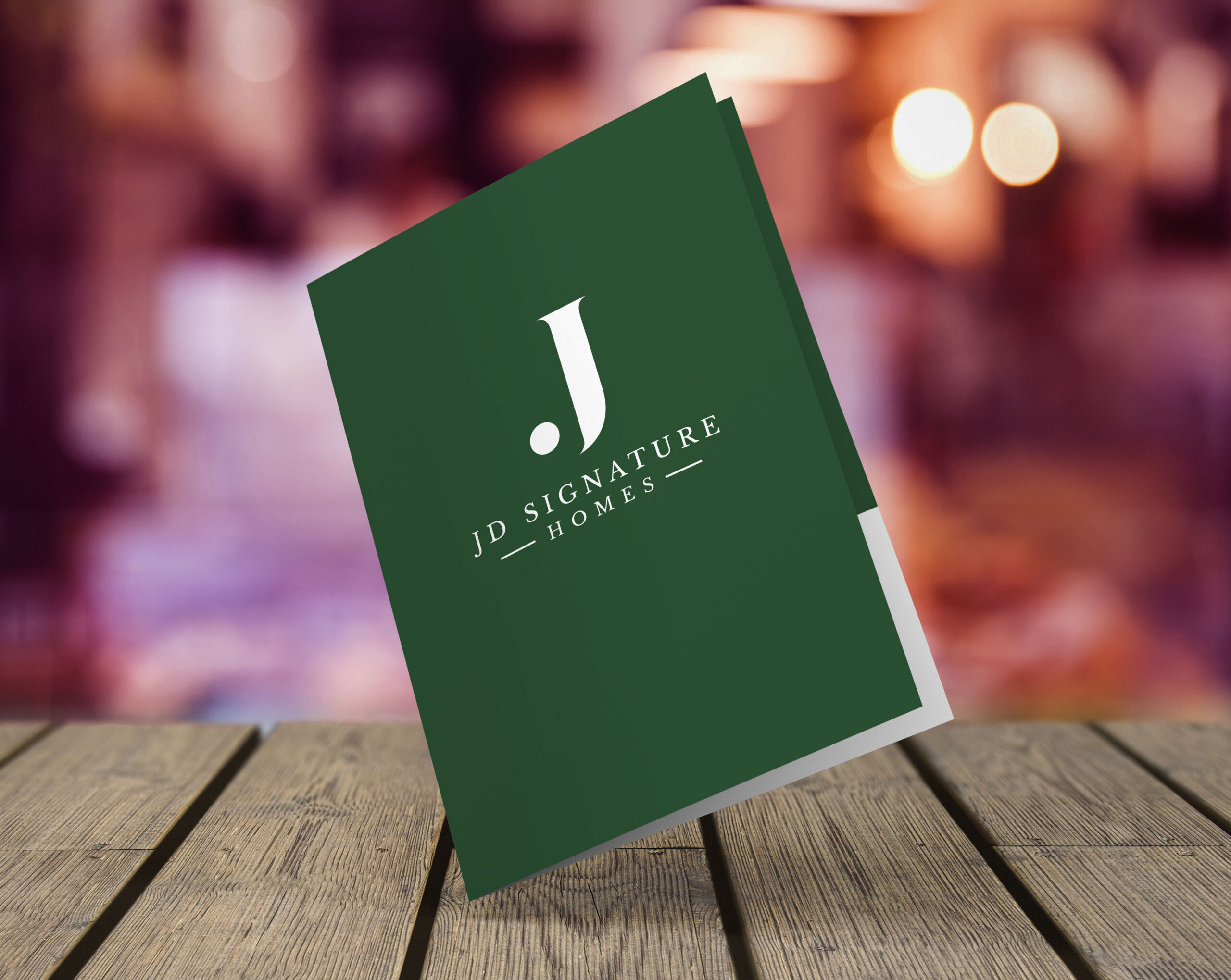 A green folder with a white J and the text JD Signature Homes stands upright on a wooden surface, subtly reflecting their expertise in logo design services, with a blurred, warmly-lit background.