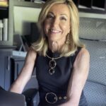A smiling woman with blonde hair, wearing a sleeveless black dress and gold jewelry, sits at her desk in a modern office, ready to discuss custom logo design services with clients. Shelves and a computer can be seen in the background.
