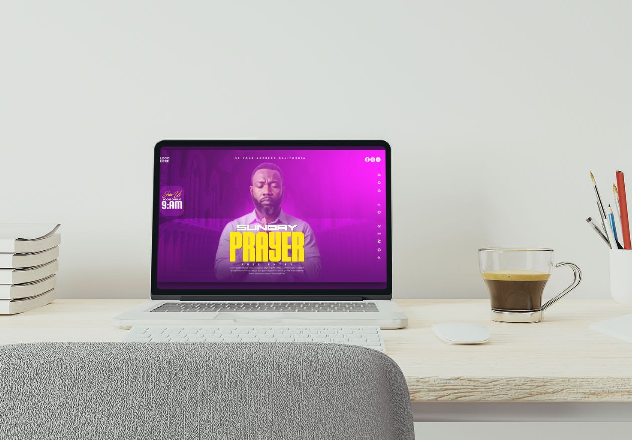 A laptop on a desk displays a vibrant custom website design for a Sunday prayer event, featuring a man with folded hands. The desk also holds stacked papers, a cup of coffee, pencils, and a gray chair.