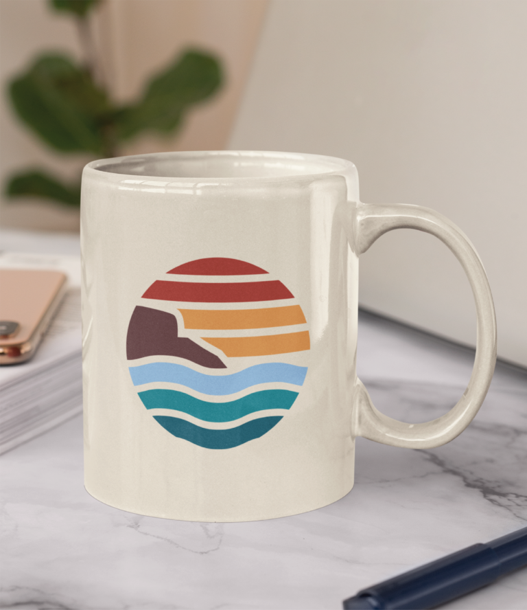 A white ceramic mug with a colorful, minimalist sunset design featuring waves, a cliff, and sun rays sits on a marble surface near a phone, notebook, and pen—an inspiring desk accessory for late-night accounting support in New Hampshire.