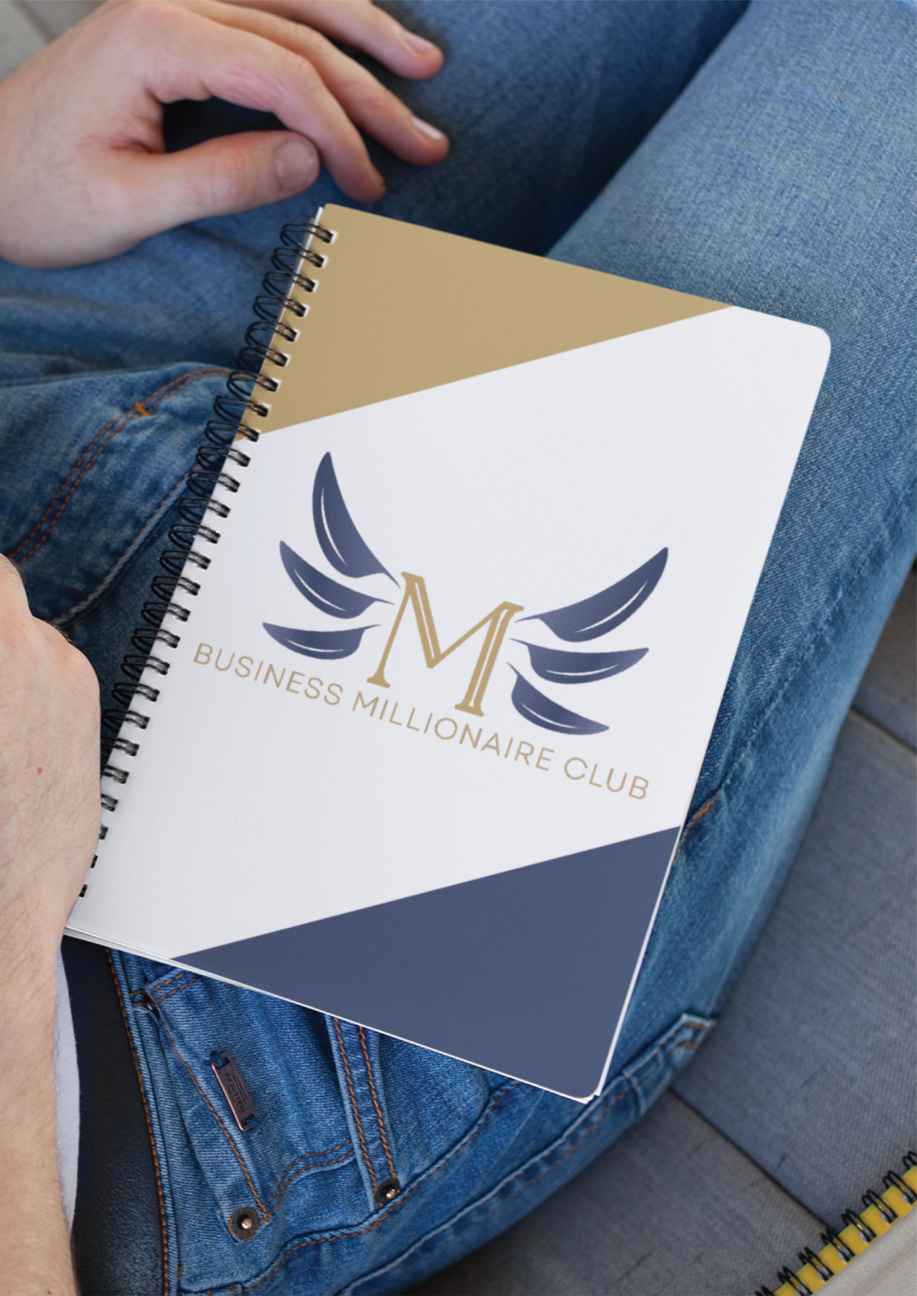 A person in jeans holds a spiral notebook with a modern design and the "M Business Millionaire Club" logo featuring stylized wings—perfect for educational consultants or anyone focused on branding.
