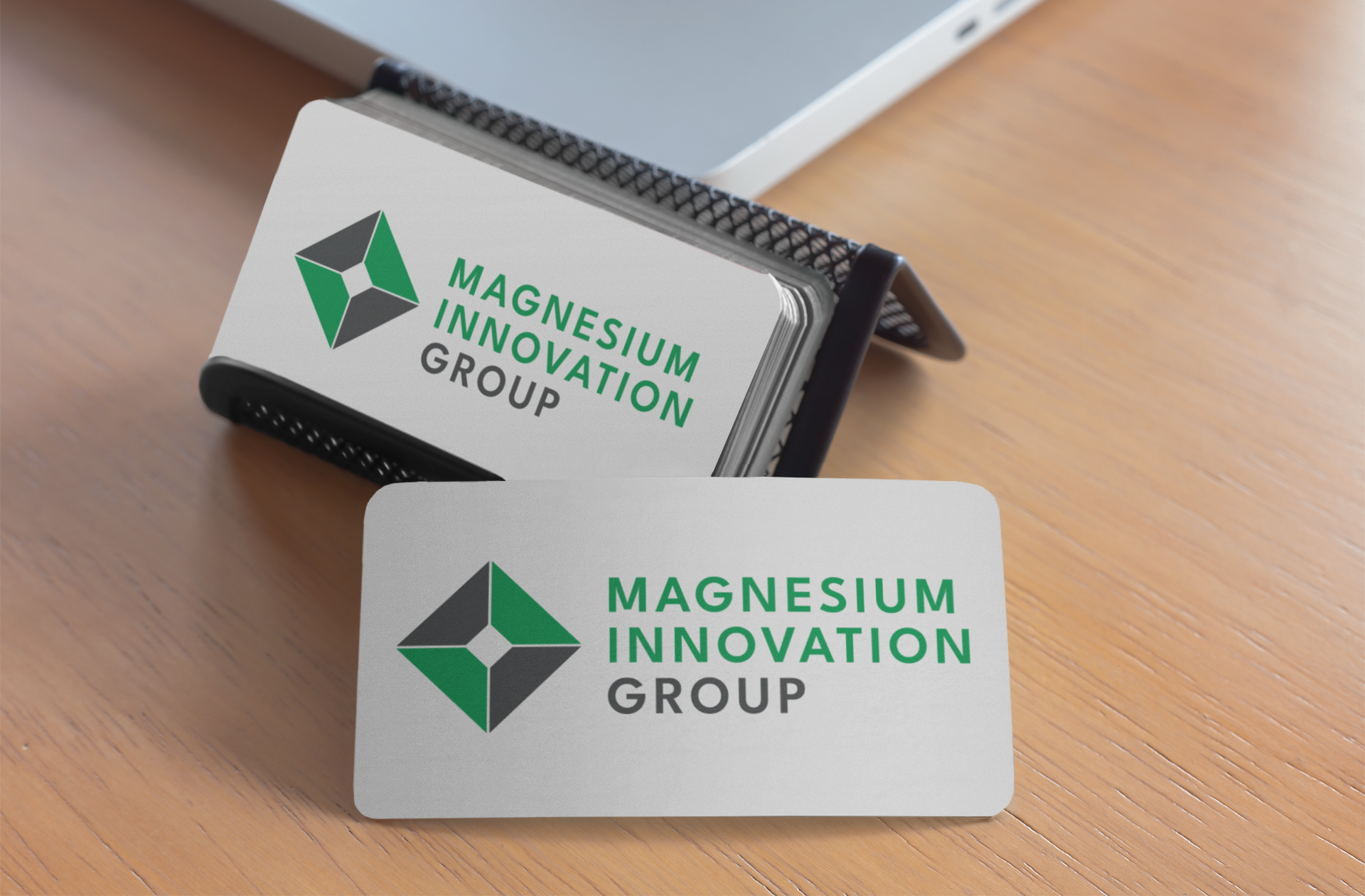 A stack of business cards in a holder and one card on a wooden desk, displaying the logo and name Magnesium Innovation Group with a green and black geometric design, highlights their expertise in Logo Design.