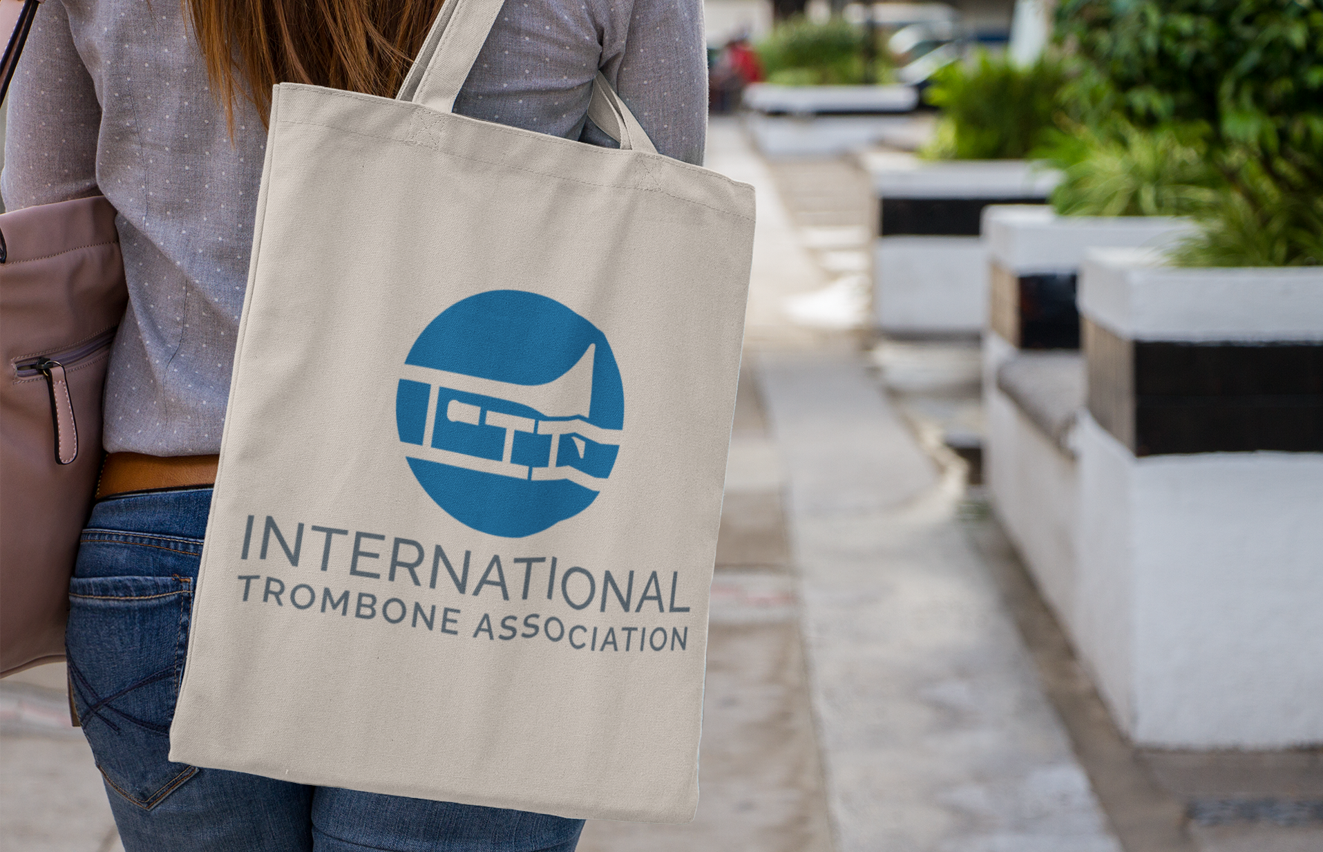 | Moonlit Media LLC A person walks outdoors in Richmond VA carrying a beige tote bag with the blue “International Trombone Association” logo, a stylized trombone inside a circle—a great example of eye-catching logo design.
