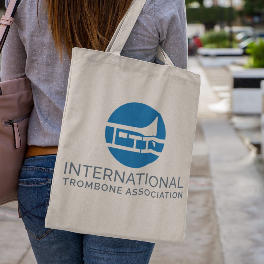 A person with long hair carries a beige tote bag over their shoulder. The bag features a blue logo designed for the International Trombone Association, showcasing creative logo design inspired by musical arts Richmond VA. The person is walking outdoors.