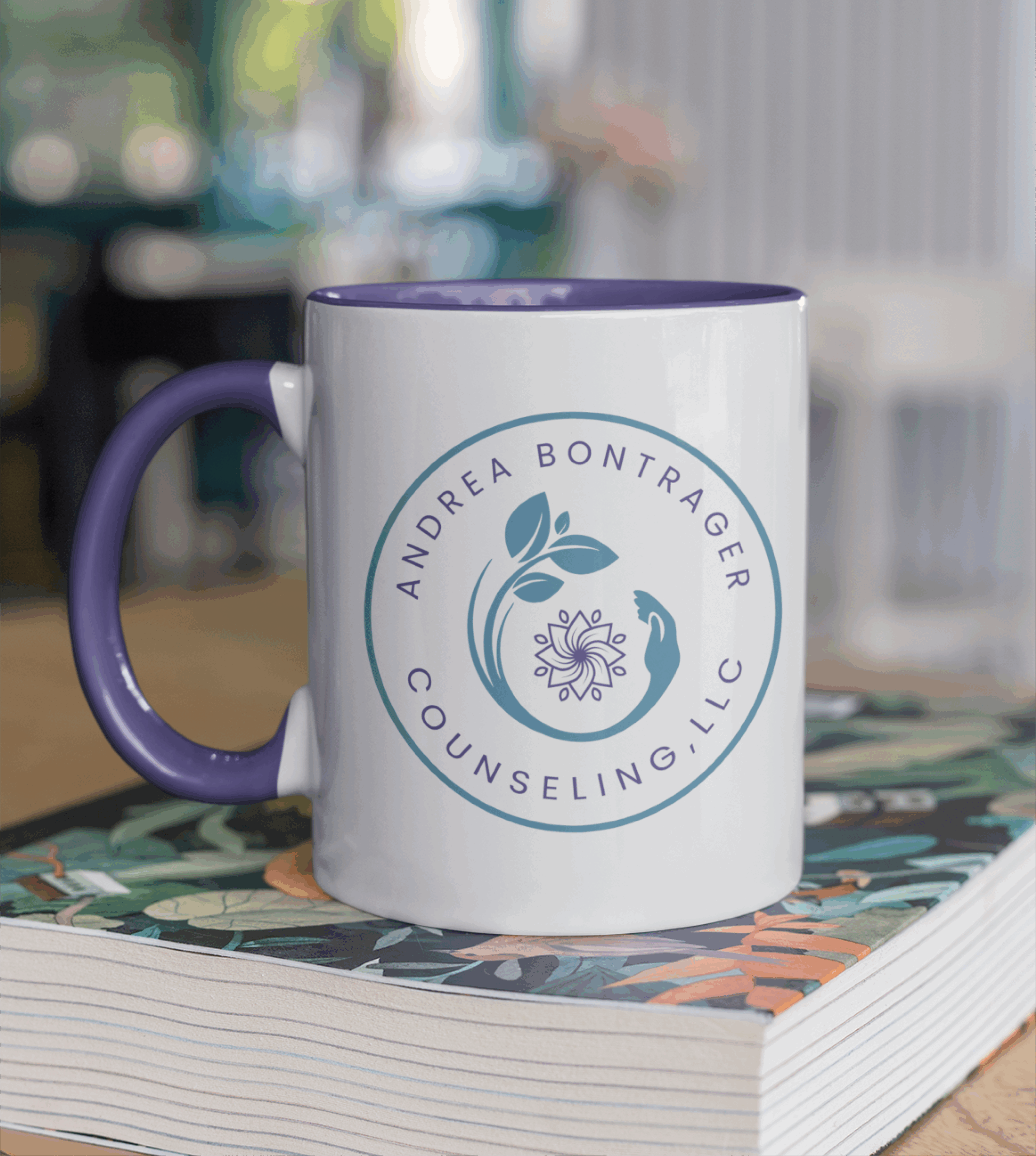 A white mug with a purple handle sits on a closed book. The mug displays the logo design for Andrea Bontrager Counseling, LLC, featuring a leaf, bird, and stylized flower within a circular motif.
