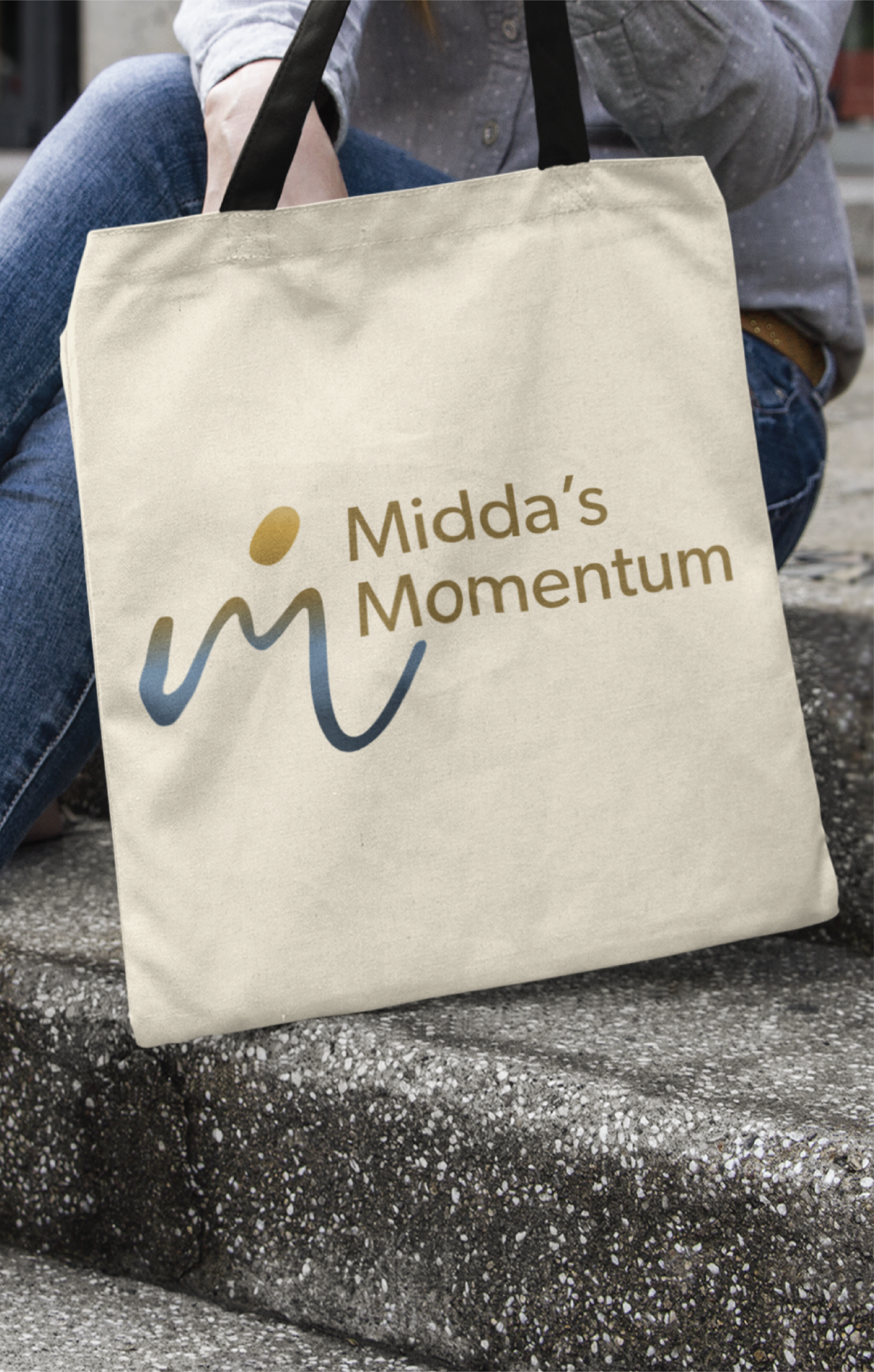 A person sitting on outdoor steps holds a beige tote bag with the Middas Momentum logo, showcasing gold and blue text and a flowing abstract design—perfect branding inspiration for educational consultants or website design professionals.