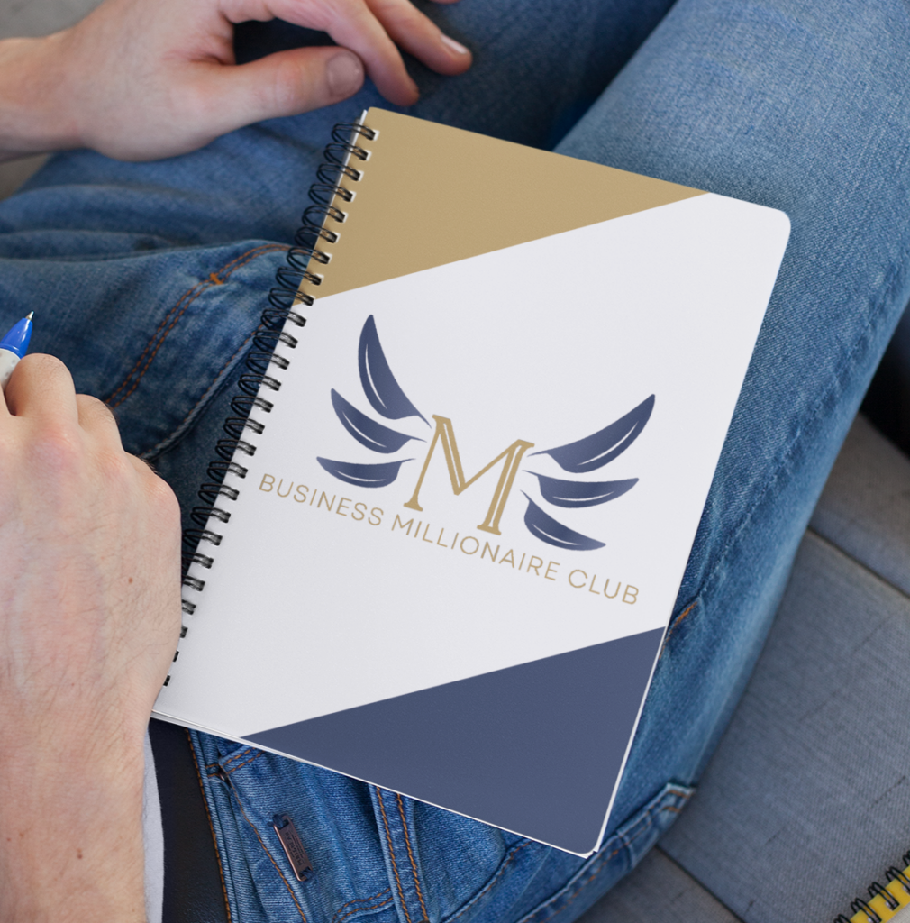 A person in jeans holds a spiral notebook with a gold, white, and navy cover that reads M Business Millionaire Club, featuring a stylized winged M logo—perfect inspiration for business consultants focused on branding.