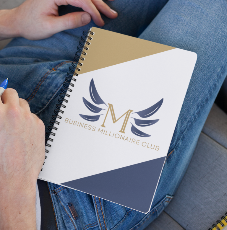 A person in jeans holds a spiral notebook with a gold, white, and navy cover that reads M Business Millionaire Club, featuring a stylized winged M logo—perfect inspiration for business consultants focused on branding.
