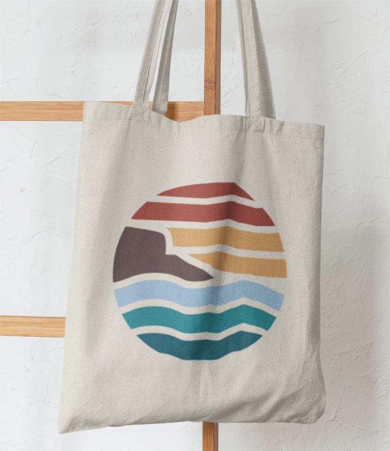 A beige tote bag with a circular design featuring stylized waves, a mountain, and a sunset in vibrant hues—reminiscent of New Hampshire’s scenic beauty—hangs on a wooden rack against a white textured wall.