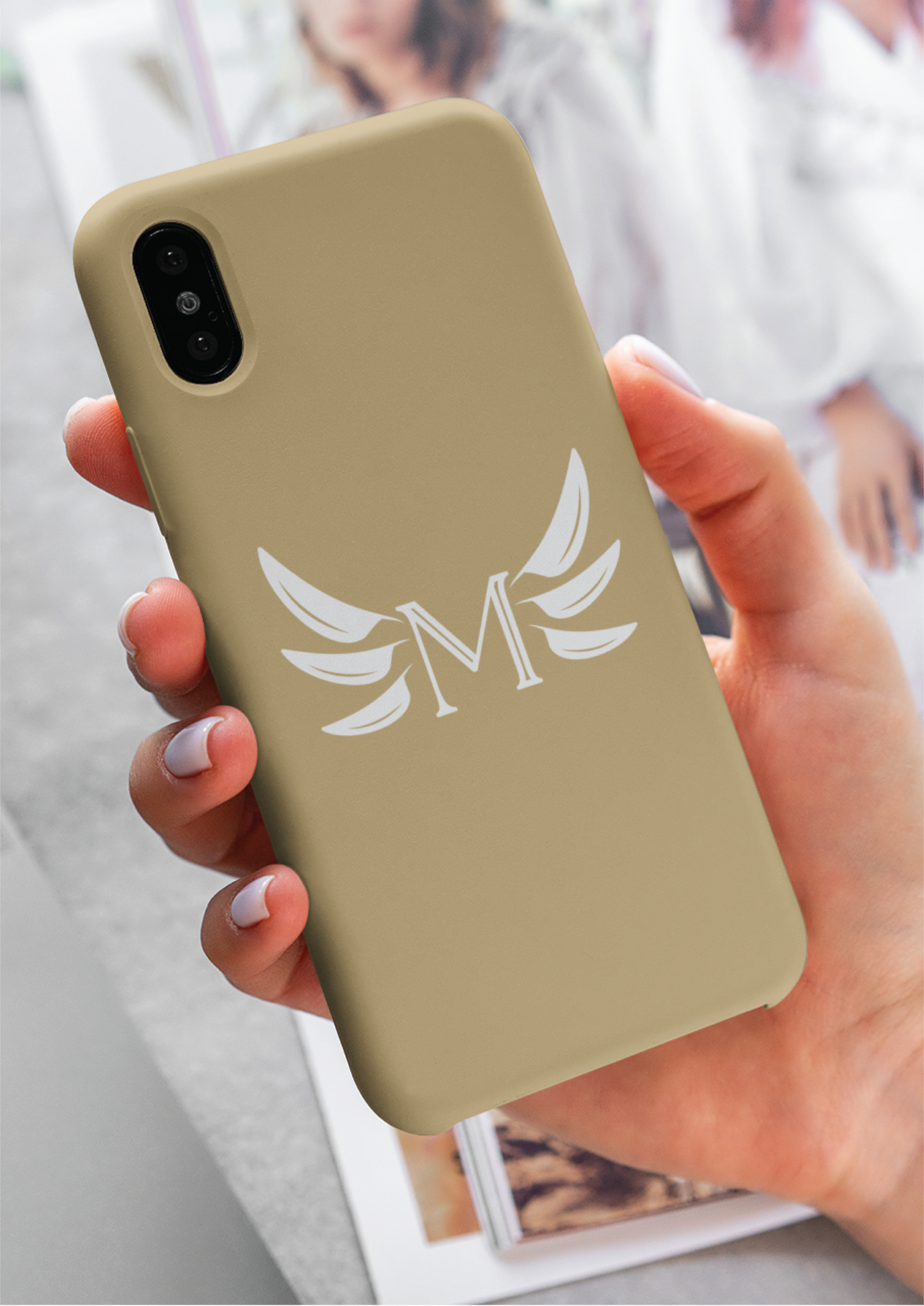 A hand holds a beige phone case featuring a white letter M with stylized wing designs, reflecting sleek branding. In the background, a blurred magazine with images adds depth to the scene.