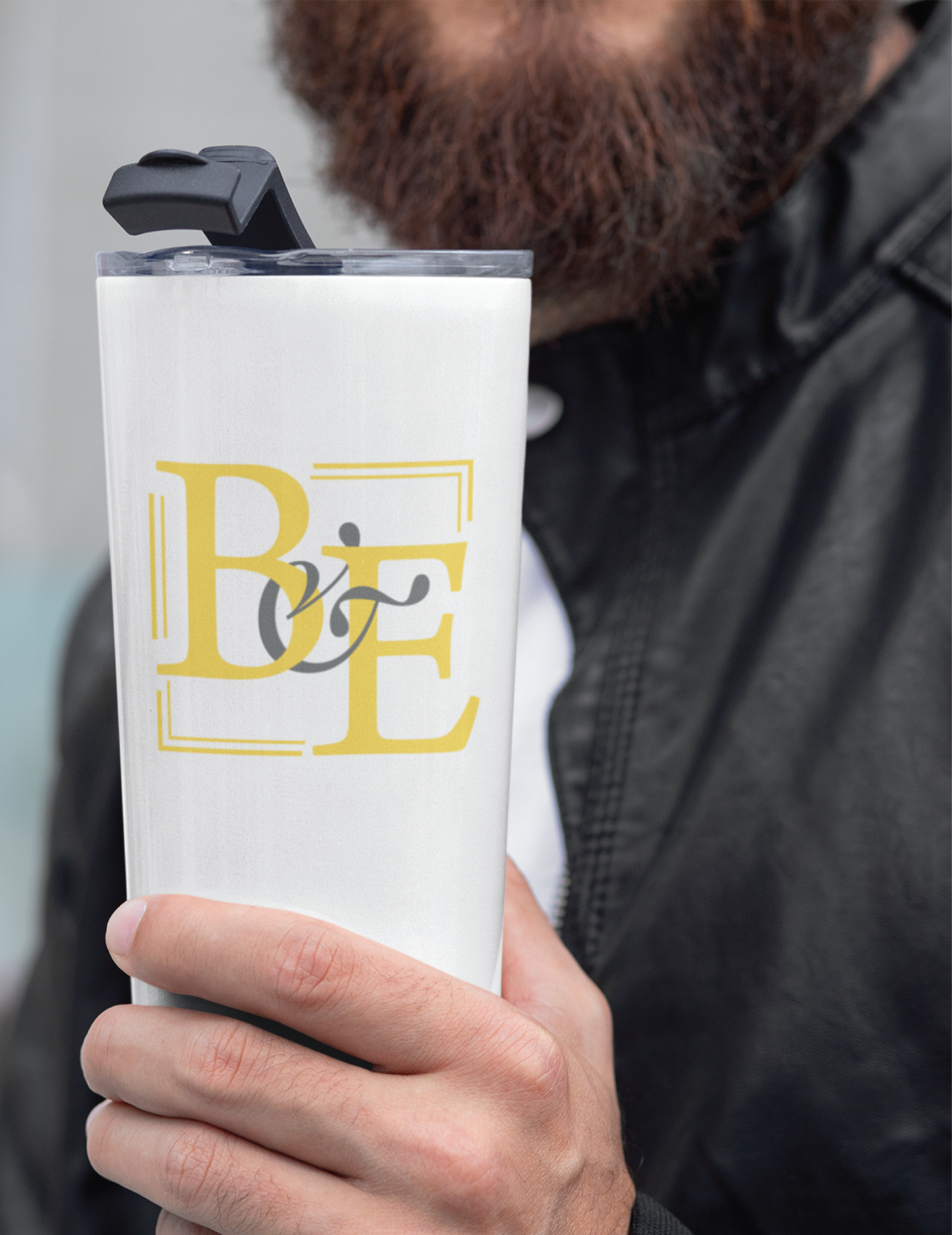 | Moonlit Media LLC A person with a beard and black leather jacket holds a white travel tumbler featuring a yellow B E logo with a bicycle graphic, showcasing unique branding that promotes wellness on the go.