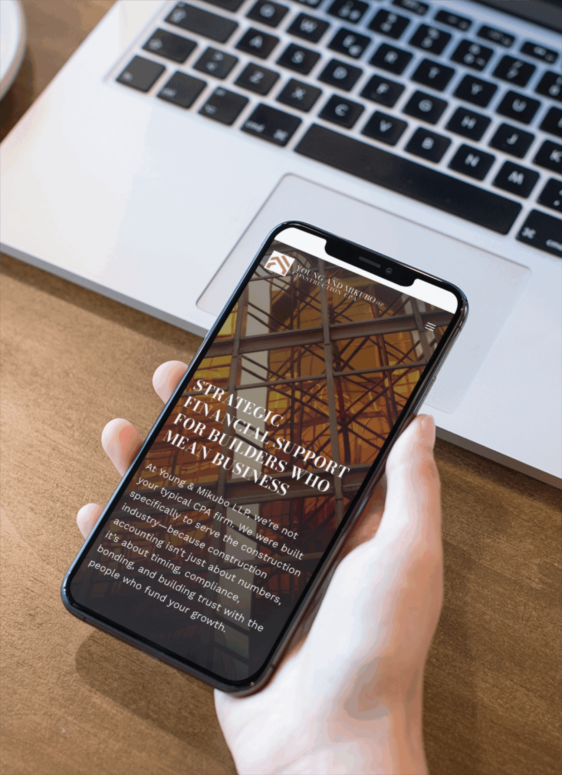 A person holds a smartphone displaying a website with text about strategic financial support for business and modern website design, featuring an abstract background with intersecting beams. A laptop keyboard is visible on the wooden table beneath the phone.